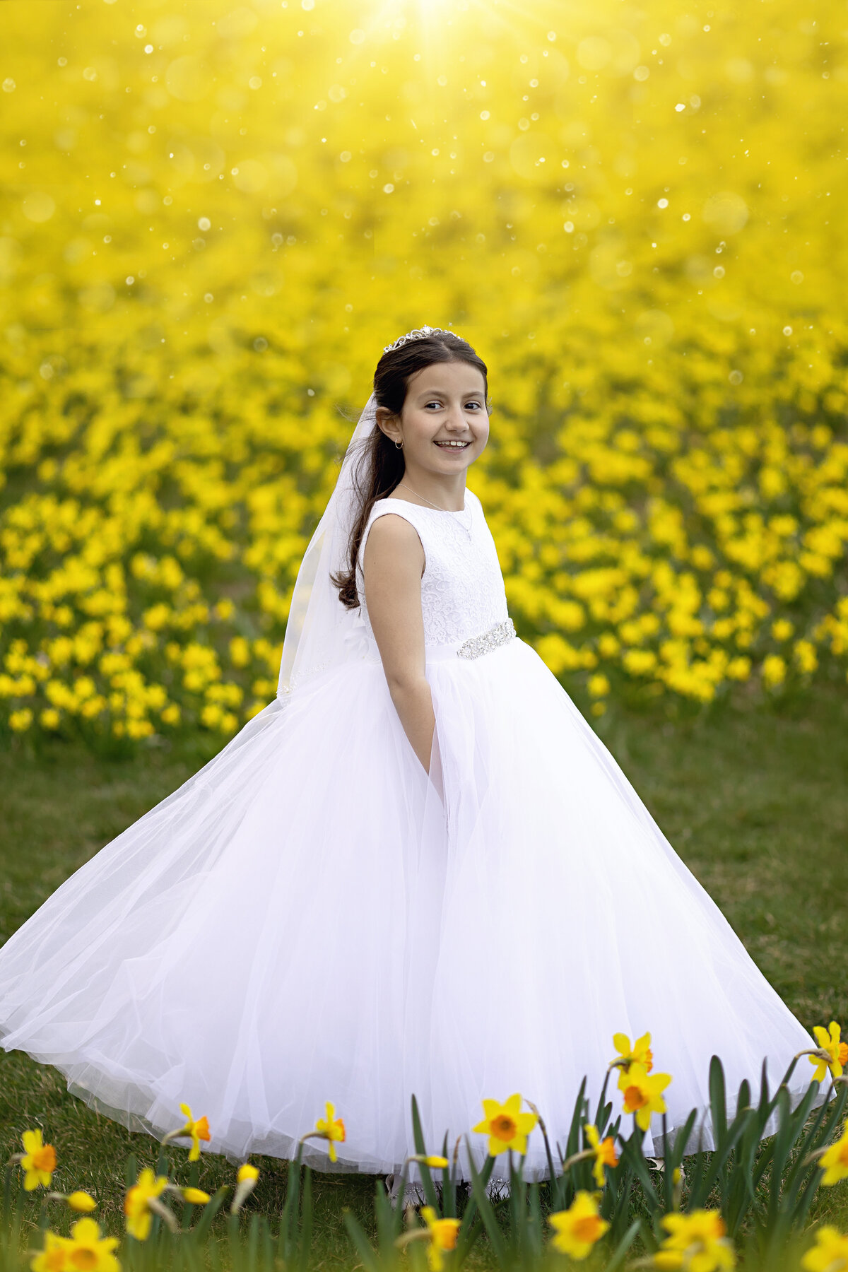 A young girl in a large white dress smiles while walking through a field of yellow daffodils