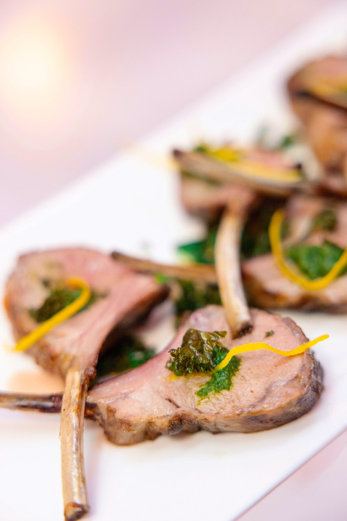 A close-up of a plate with several pieces of cooked lamb chops garnished with a green herb topping and thin lemon zest strips, arranged artfully on a white rectangular dish.