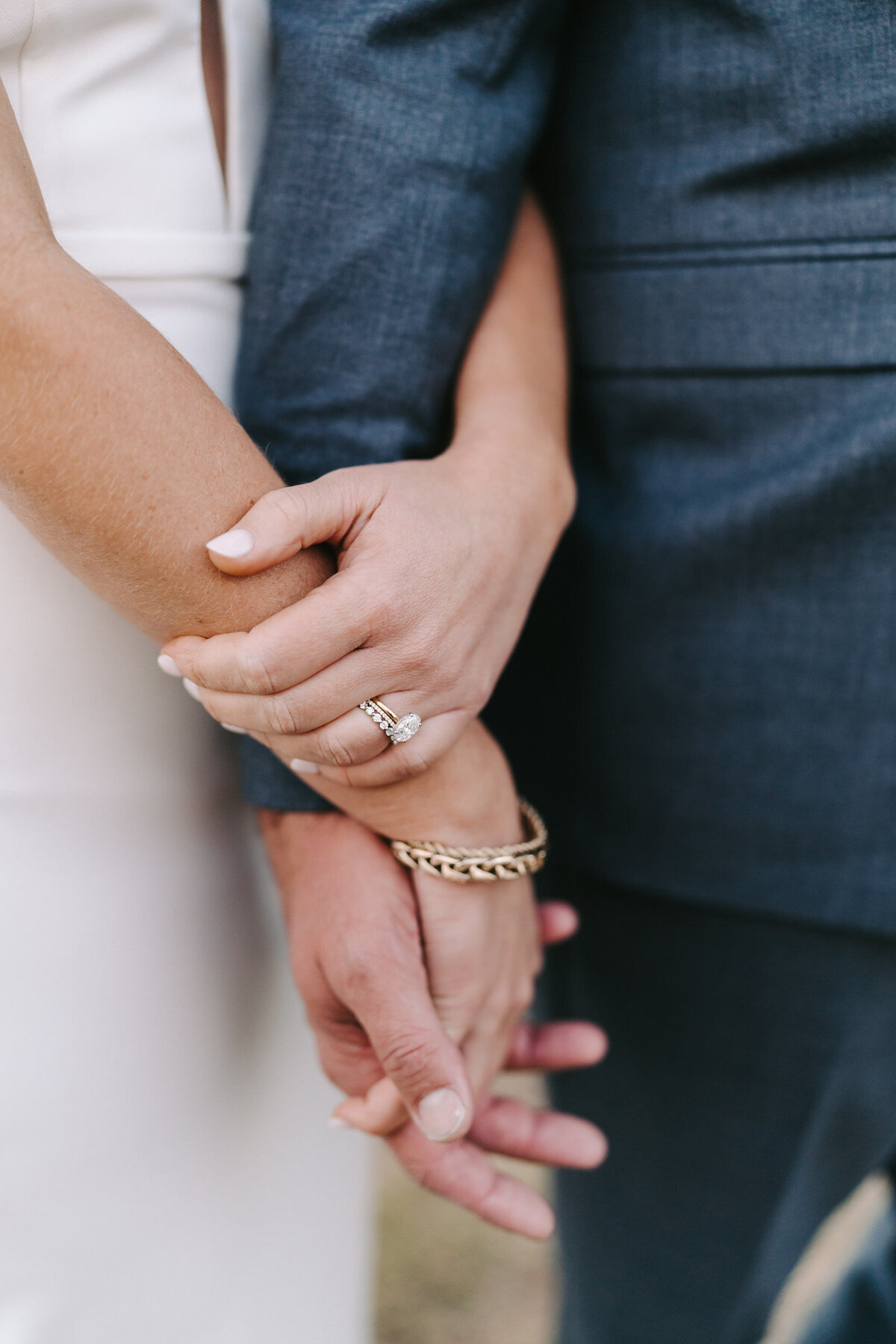 A colorful photograph of Carly and Bradley on their wedding day at Morgan Creek Barn in Dripping Springs, near Austin, Texas. The photo is a close-up of their clasped hands showing her wedding ring and wedding band along with her gold bracelet. Wedding photography taken by Stacie McChesney/Vitae Weddings.