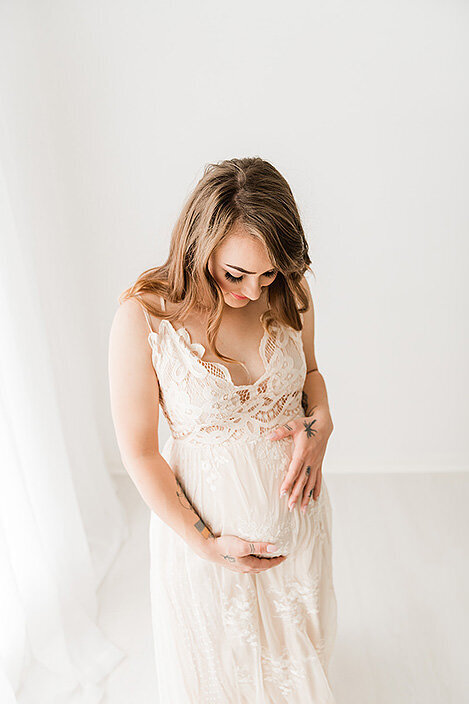 A tender moment as an expecting mother in a lace gown looks down at her baby bump in a bright white studio.