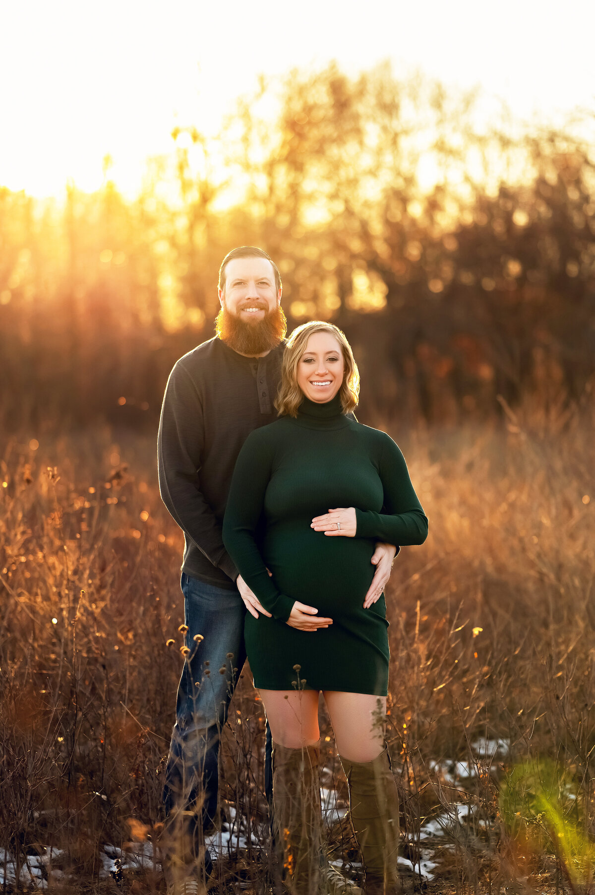 Expecting couple standing in a sunlit Wisconsin field during golden hour, with the pregnant woman wearing a forest green dress and her partner embracing her from behind.