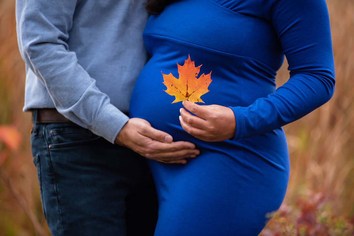 up close photo of pregnant woman and her husband cradling her belly and her holding a maple leaf for their fall maternity photography session.
