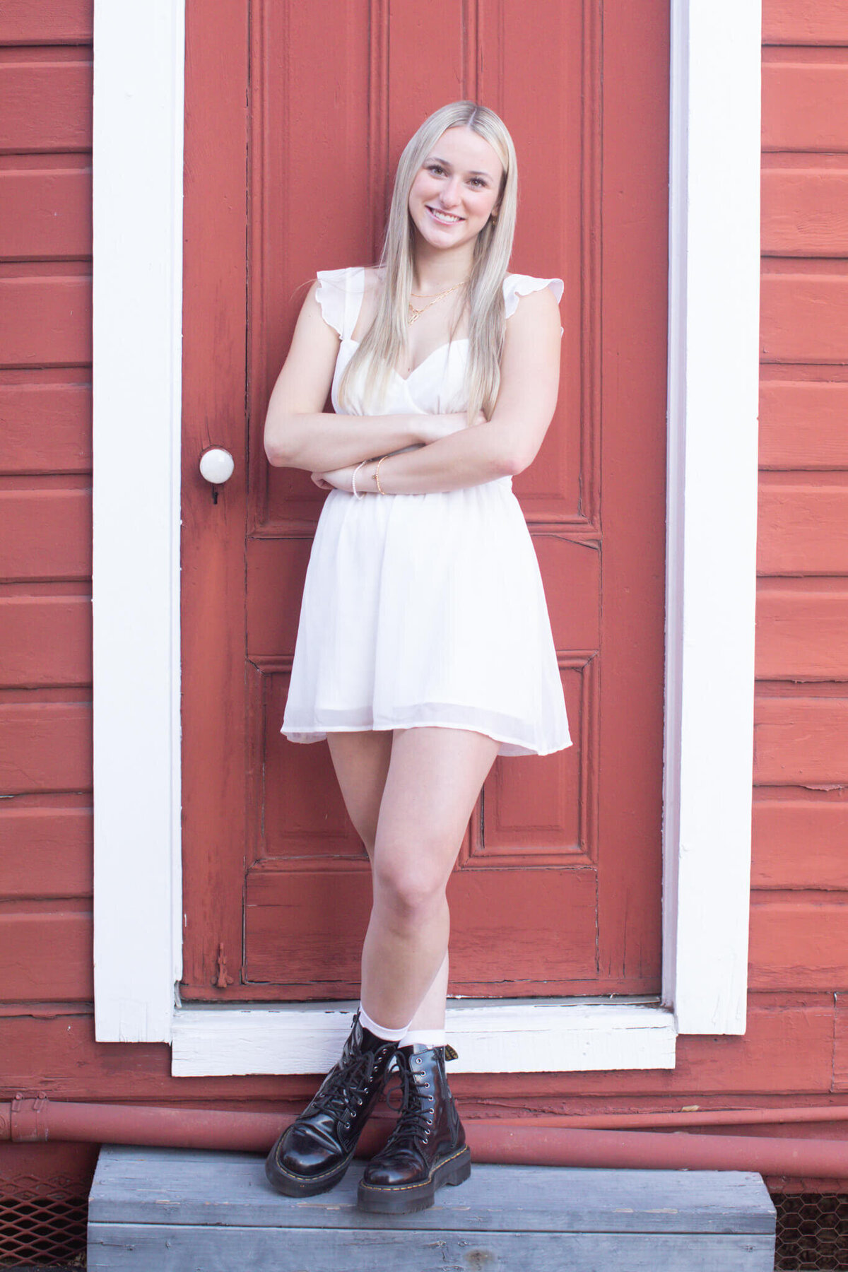 A young woman with long blonde hair stands smiling in front of a red wooden door. She is wearing a white dress and black boots, with her arms crossed. The door has white trim, and the background is a red paneled wall.