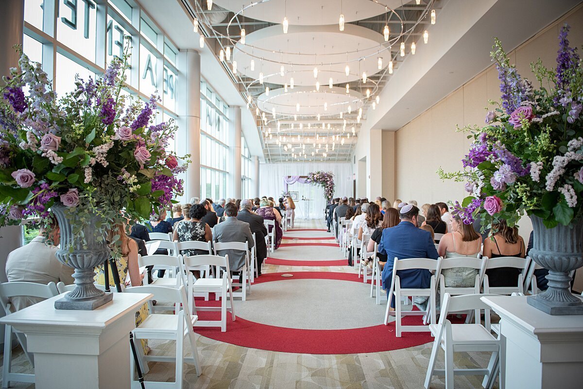 A wedding ceremony set with white  garden chairs on red and beige carpet. On either side of the aisle are two  rectangular white pillars, two grey stone urns with  large purple floral arrangements with pink roses, purple roses, white stock and purple hydrangea. At the end of the aisle is a wedding arbor covered with greenery, purple roses, pink roses, white stock, blush roses, and purple hydrangea.