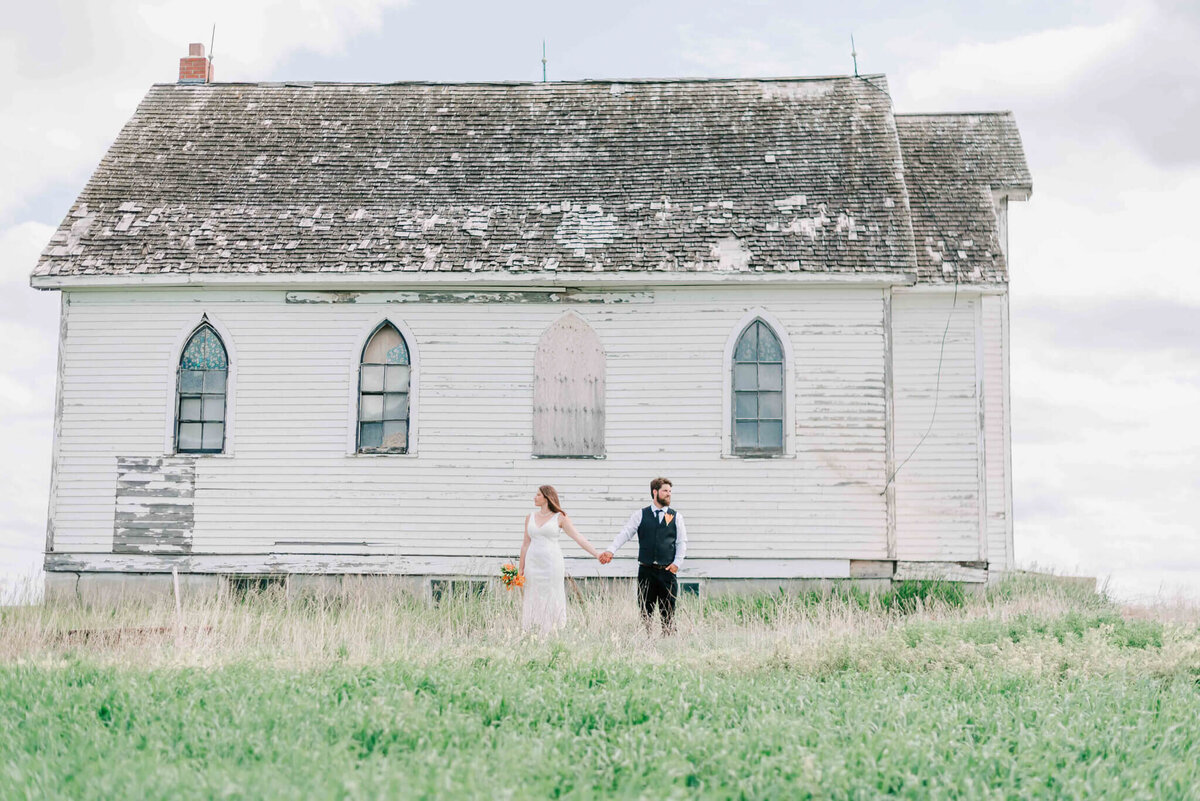 Bride and groom holding hands pulling away from each other by an abandoned church by Allen Saskatchewan.