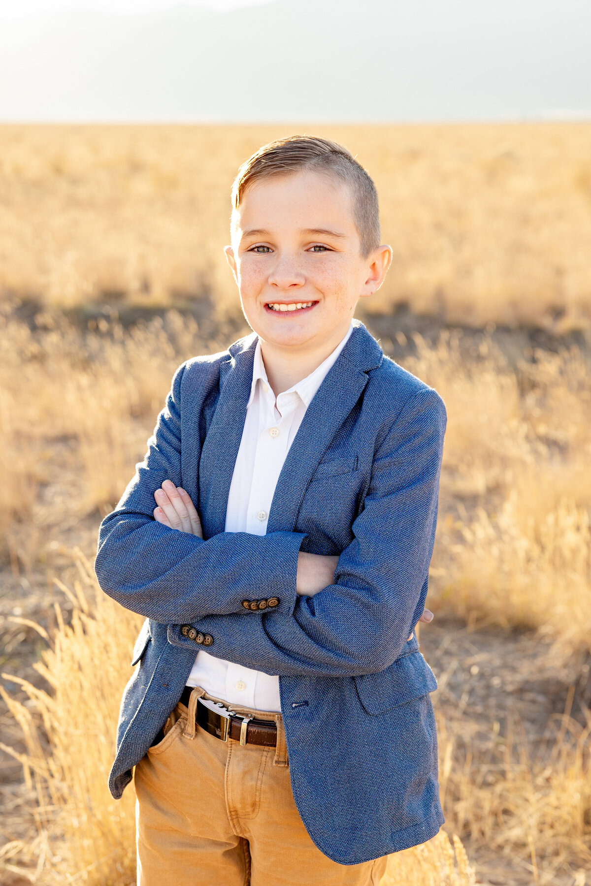 A young boy wearing a stylish blue blazer crosses his arms and smiles confidently while standing in a field during a Cassie Lee Photography session.