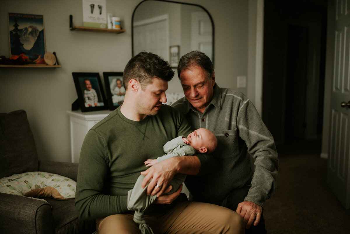 In the cozy nursery of Champlin, Minnesota, this touching image portrays a newborn baby boy cradled by his dad, with the baby's grandfather looking down at the newest addition to their family. Three generations share a moment of profound connection and love, creating a heartwarming memory to be treasured.