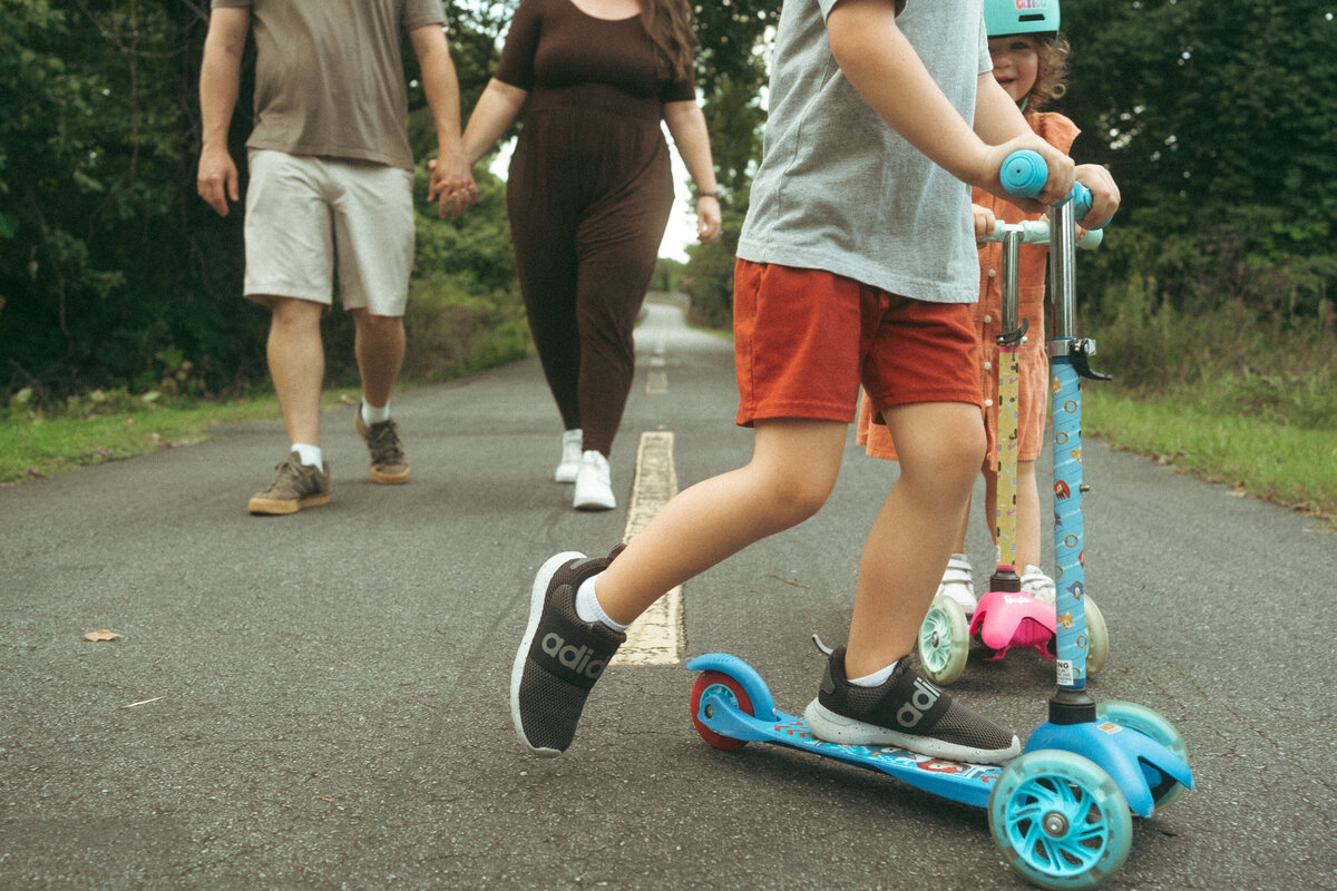 A young boy and his little sister riding schooters at Noonday Creek Trail while their parents walk behind them. 