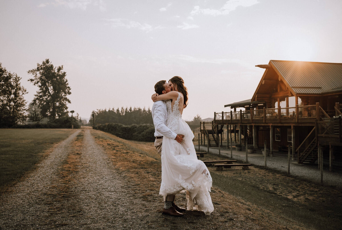 Bride and groom share a romantic kiss at sunset by a rustic barn, captured by a wedding photographer.