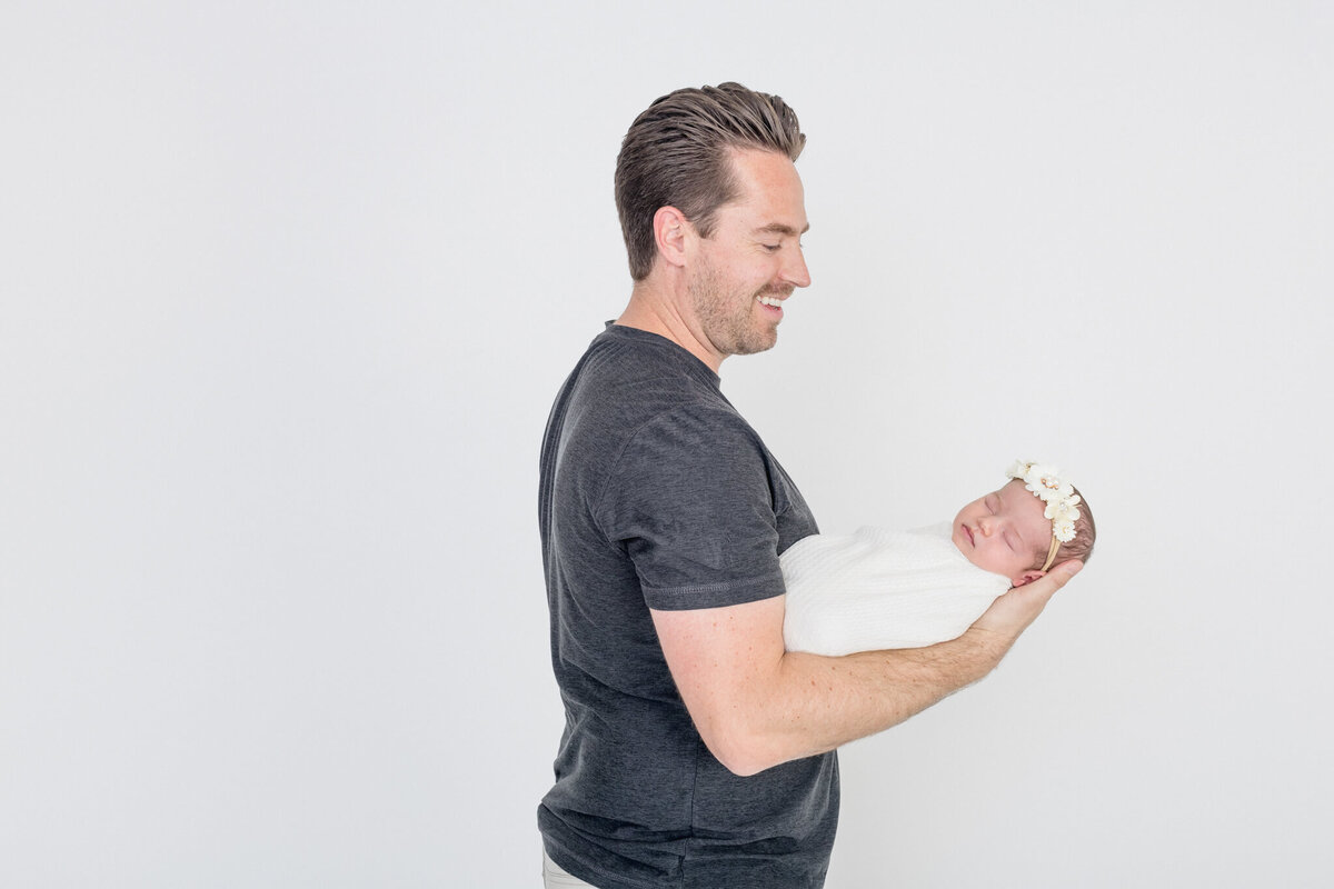 A new father with brown hair and gray shirt holding his sleeping newborn baby in one arm while gazing at her in a white studio in Tustin.