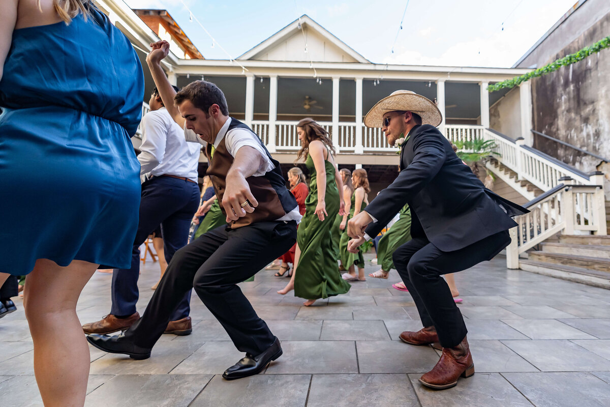 A group of people dressed in formal attire dancing enthusiastically outdoors on a tiled patio. One man wears a cowboy hat and boots while others join the fun in suits, dresses, and green gowns. A staircase is in the background.