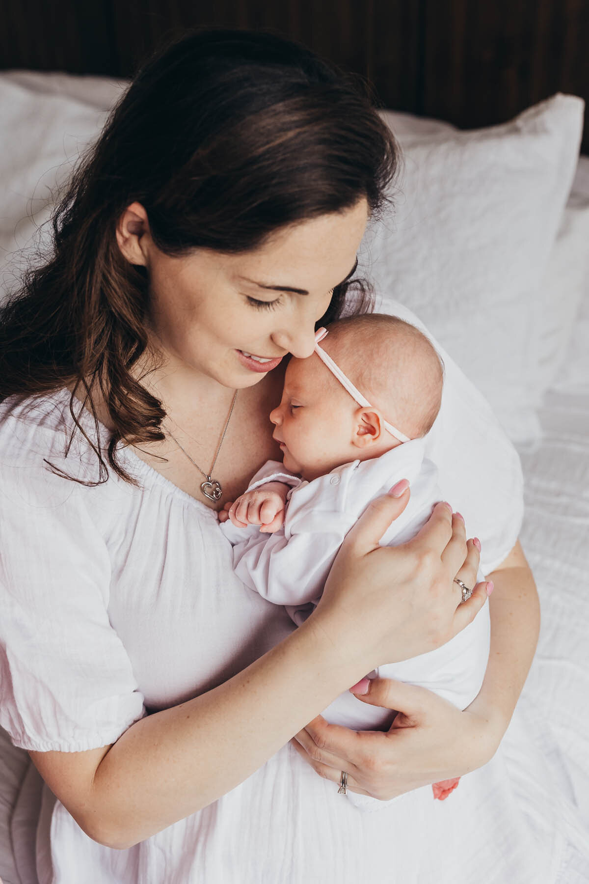 a mother and baby both wearing white sit on a bed while mom holds baby in her arms