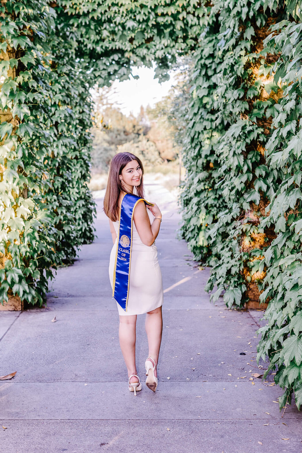 A woman in a white dress and heels stands on a path framed by lush green ivy, wearing a blue graduation stole. She is smiling and looking back over her shoulder, conveying a sense of accomplishment and celebration.