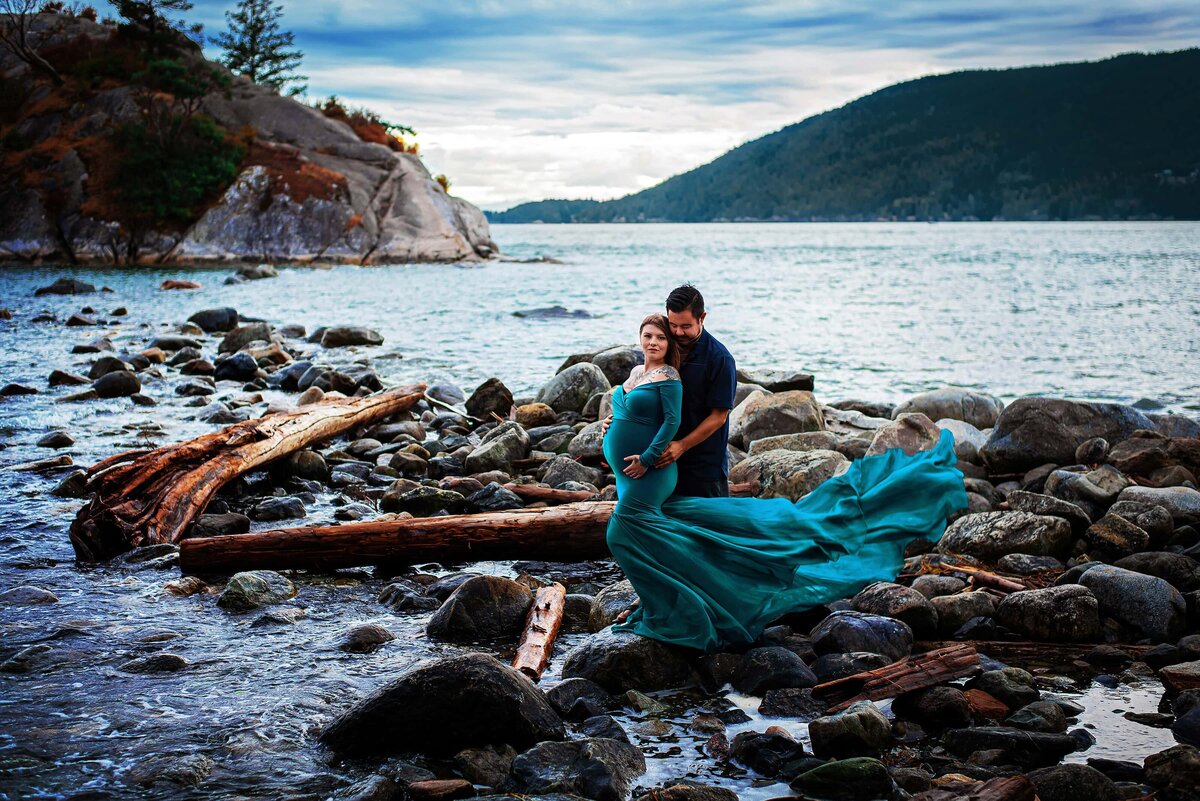 Expectant mom in teal chiffon dress with husband on rocky beach.
