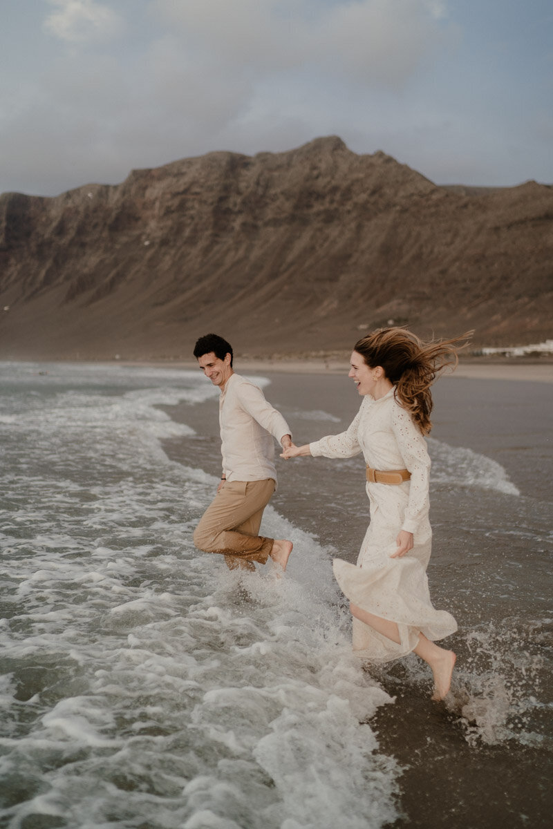 Elopement sur la plage de Famara à Lanzarote, avec un couple courant au bord de l’eau face aux falaises volcaniques.