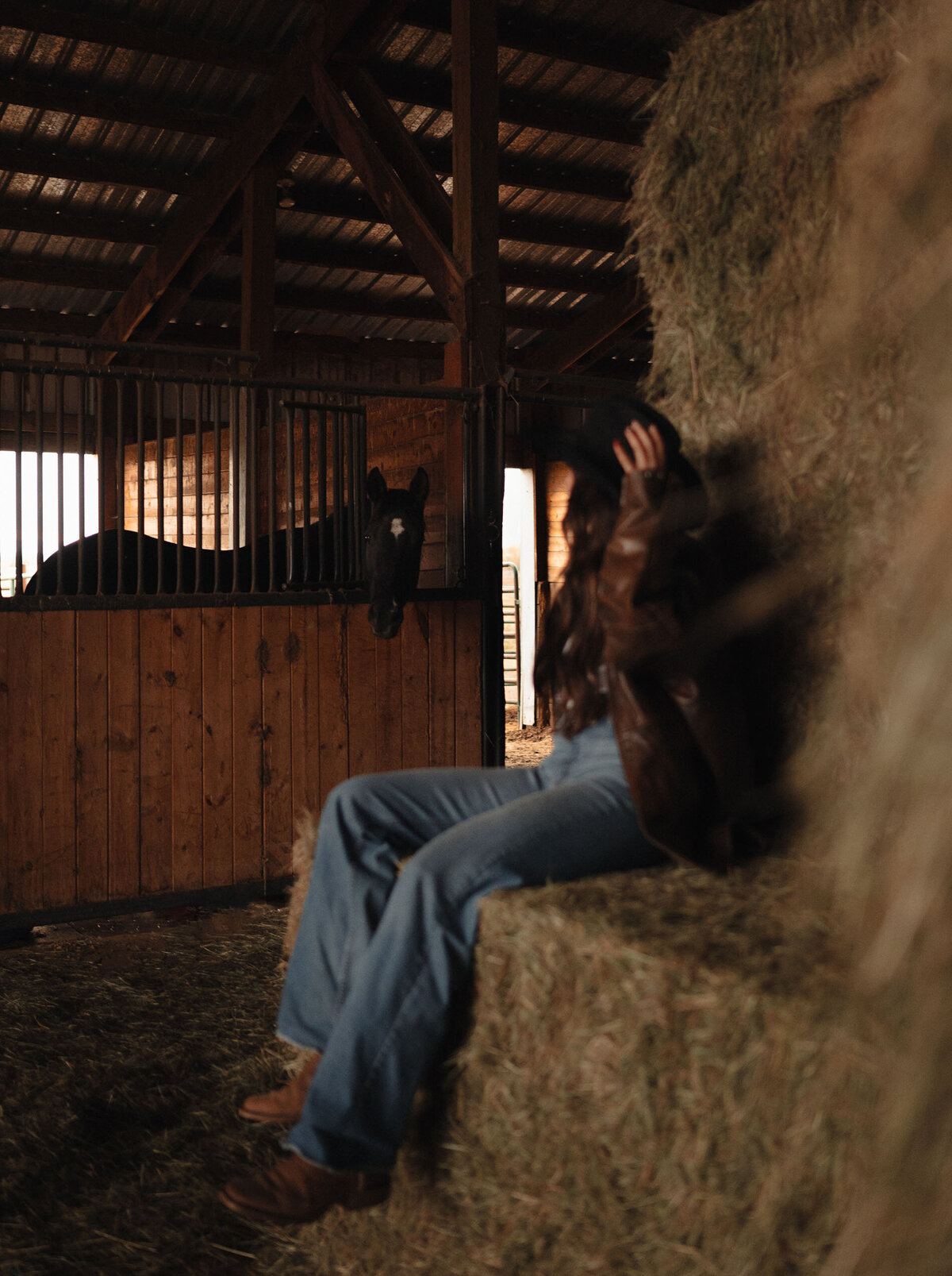 cowgirl sits on bales of hay while looking at her horse during personal brand photoshoot