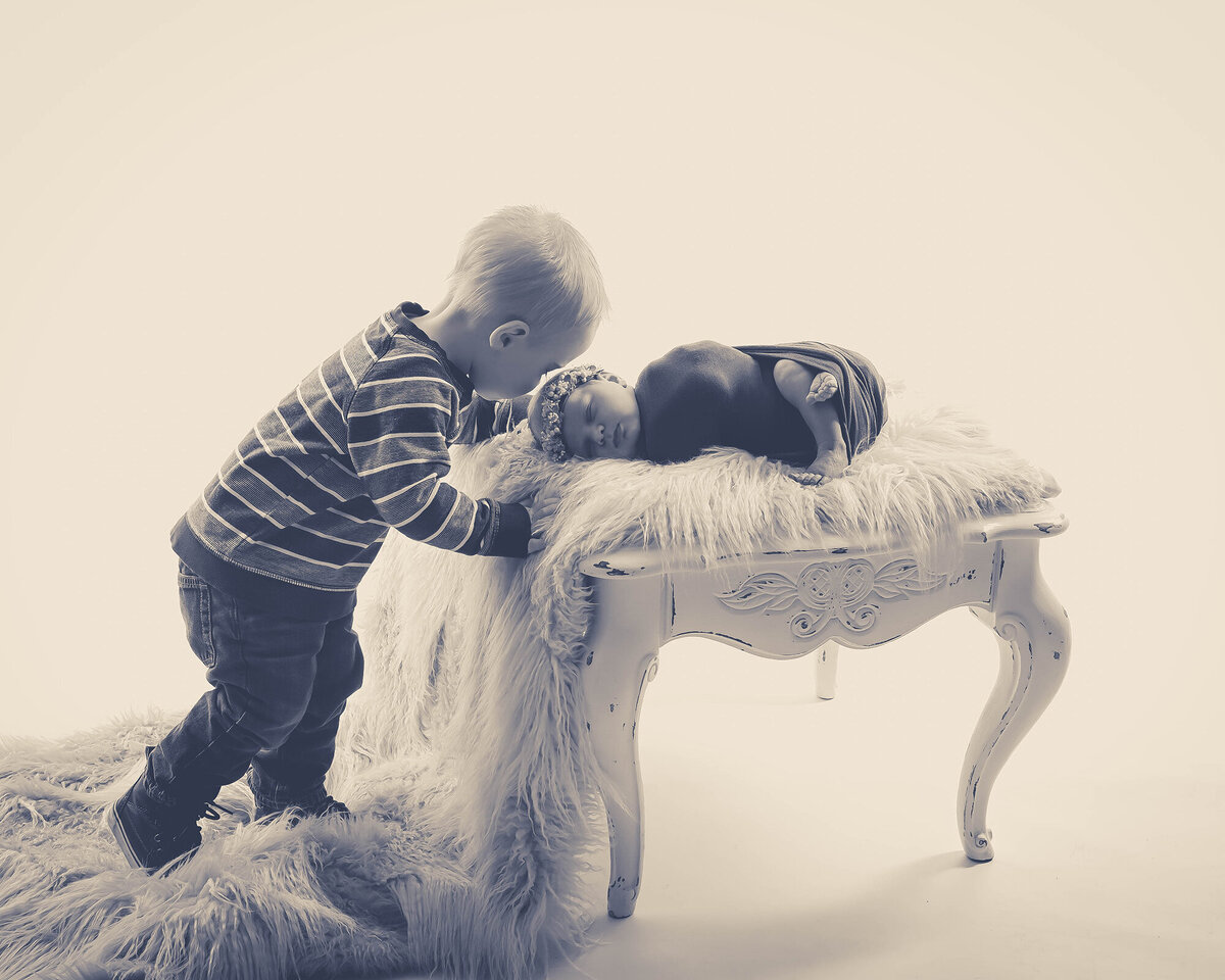 A toddler boy kisses the head of his sleeping newborn baby sister in a studio