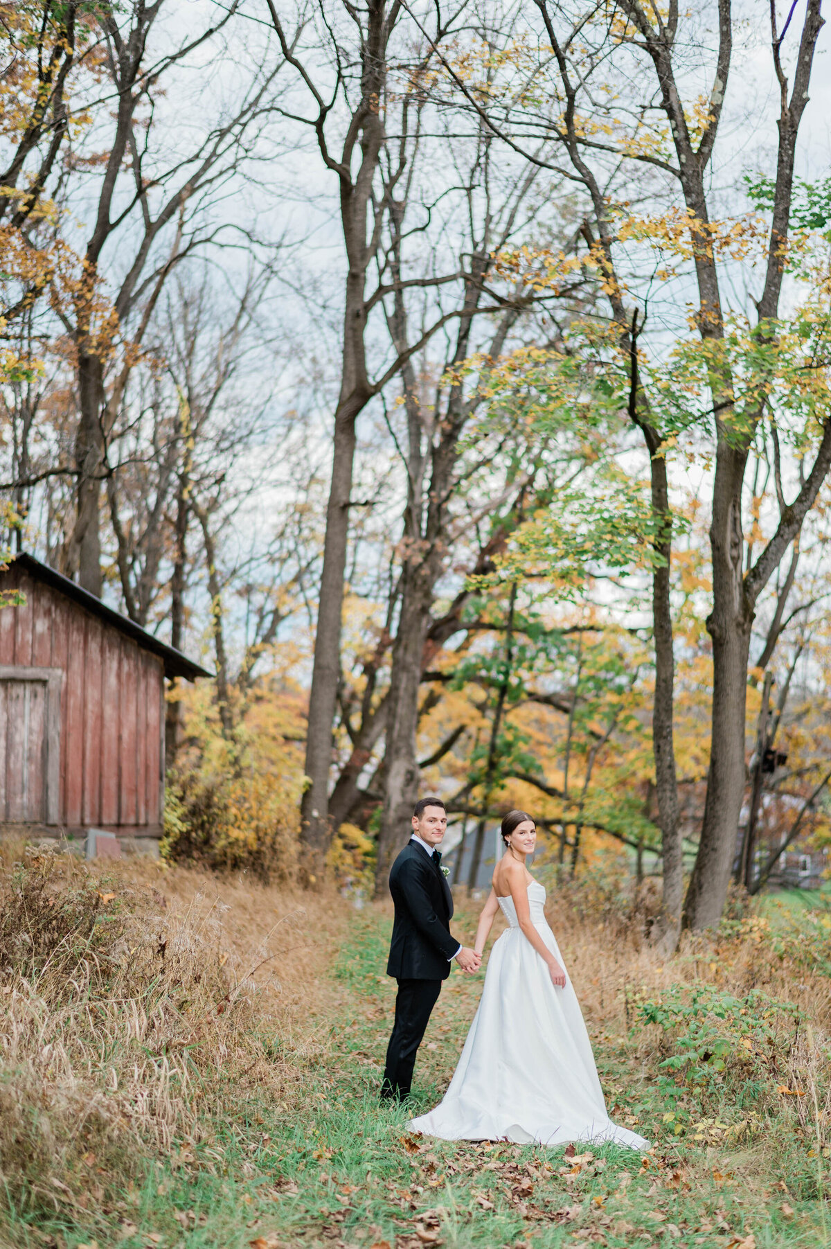 The couple stands on a grassy path near a rustic barn at Meadow Ridge on Hudson, holding hands