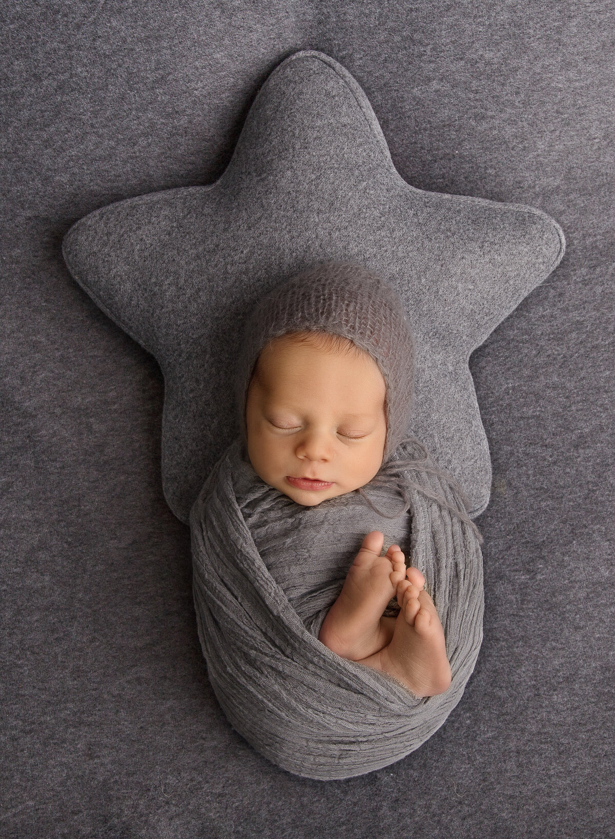 Baby boy swaddled in grey with this does peeking out is placed atop of a large grey felt star pillow for his newborn photoshoot. Captured by top Brooklyn newborn photographer Chaya Bornstein