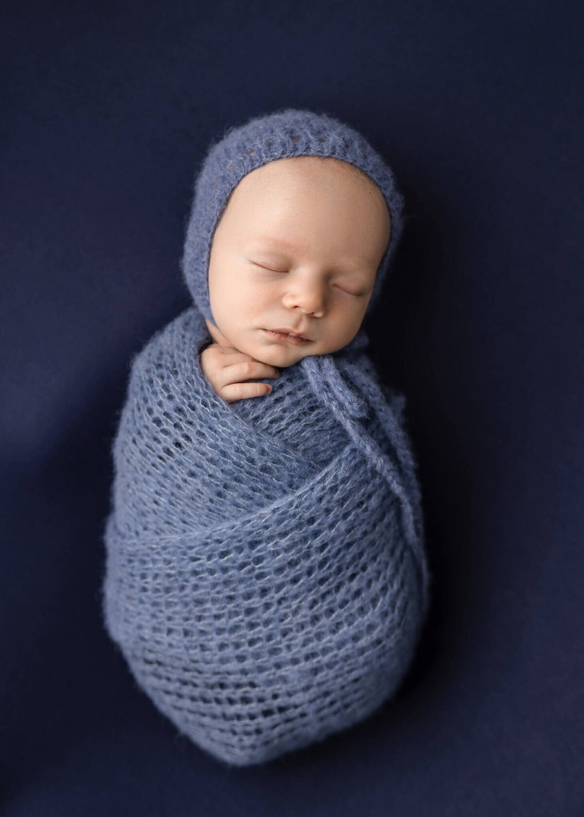 newborn baby wrapped in a blue knitted wrap and bonnet asleep on blue fabric