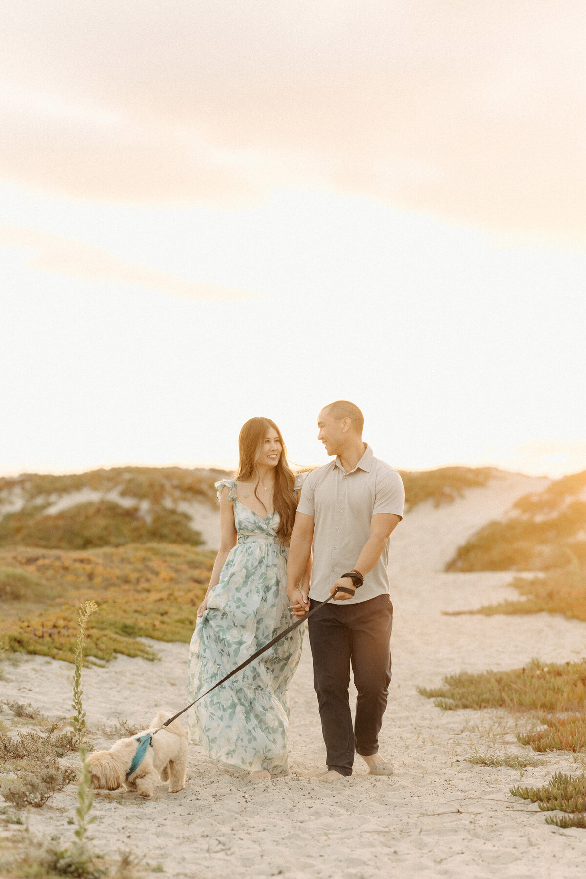 Couple walking hand in hand along the sandy paths of Coronado Beach at sunset, with their small dog on a leash.