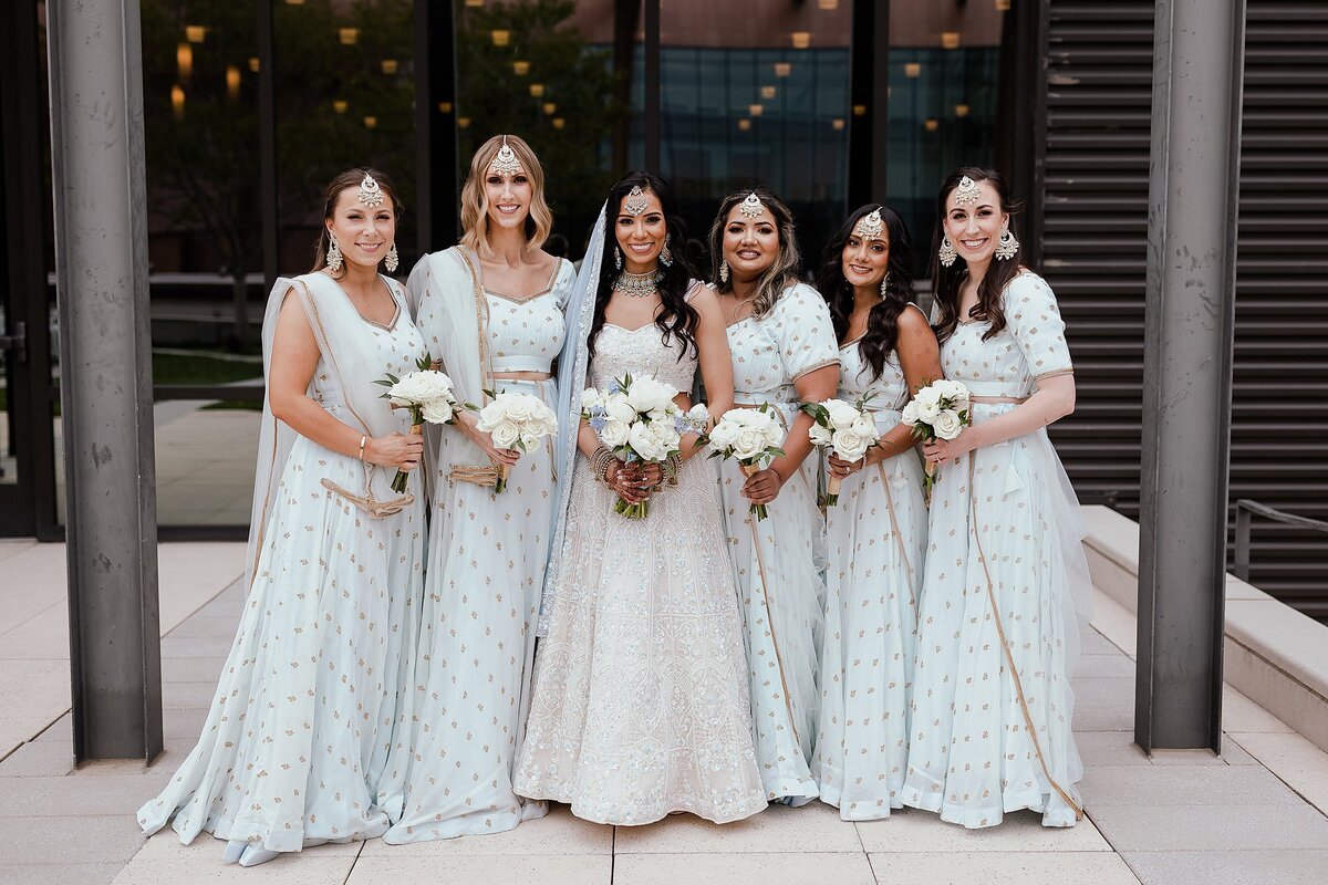 Hindu bridal party dressed in light blue sarees with gold accents standing on either side of an Indian bride wearing a white beaded saree with a light blue dupatta. The Nashville bridal party are all holding white floral bouquets.