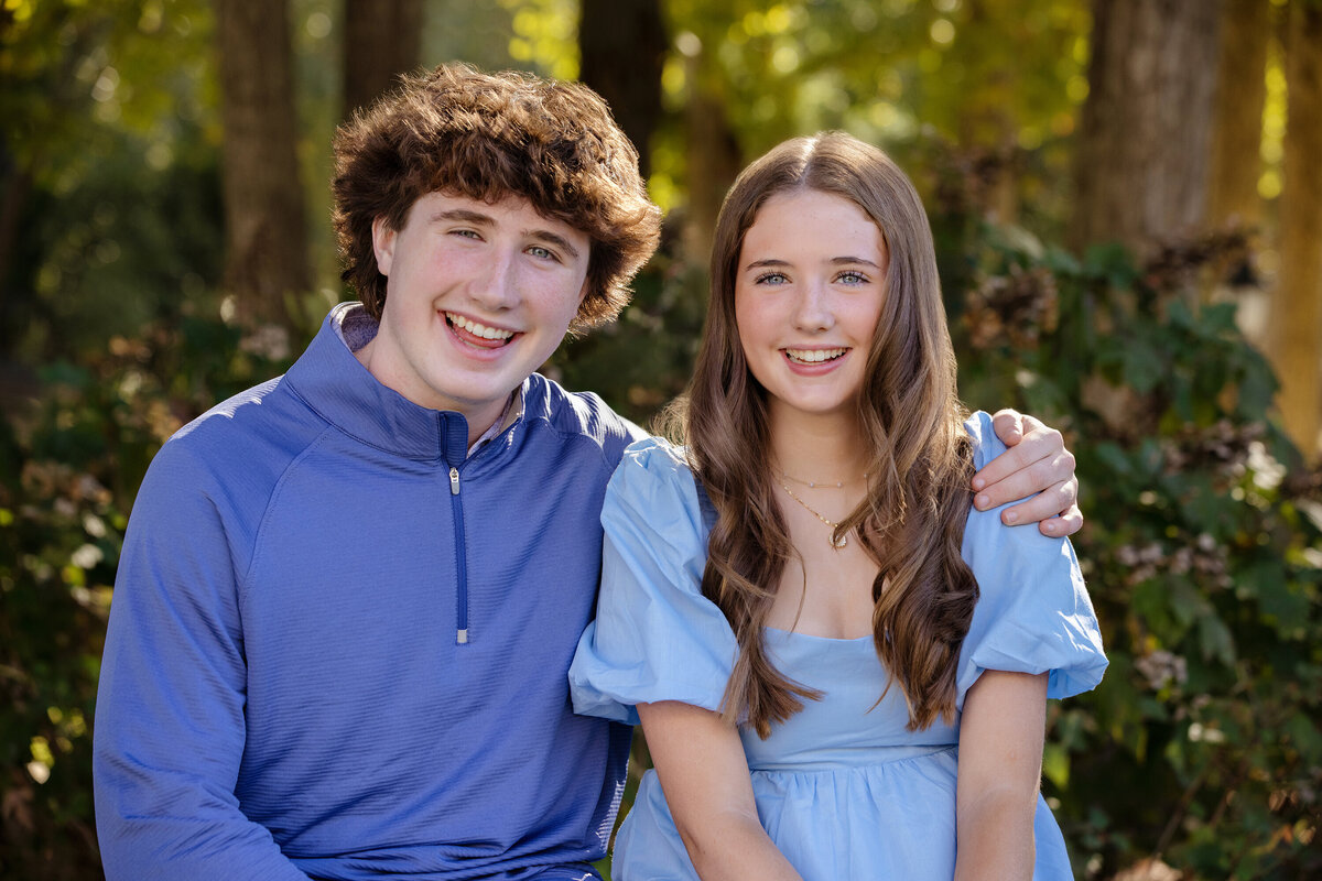 Sibling portrait of brother and sister sitting close on a garden bench with autumn greenery in Ballantyne NC, a calm, connected moment.
