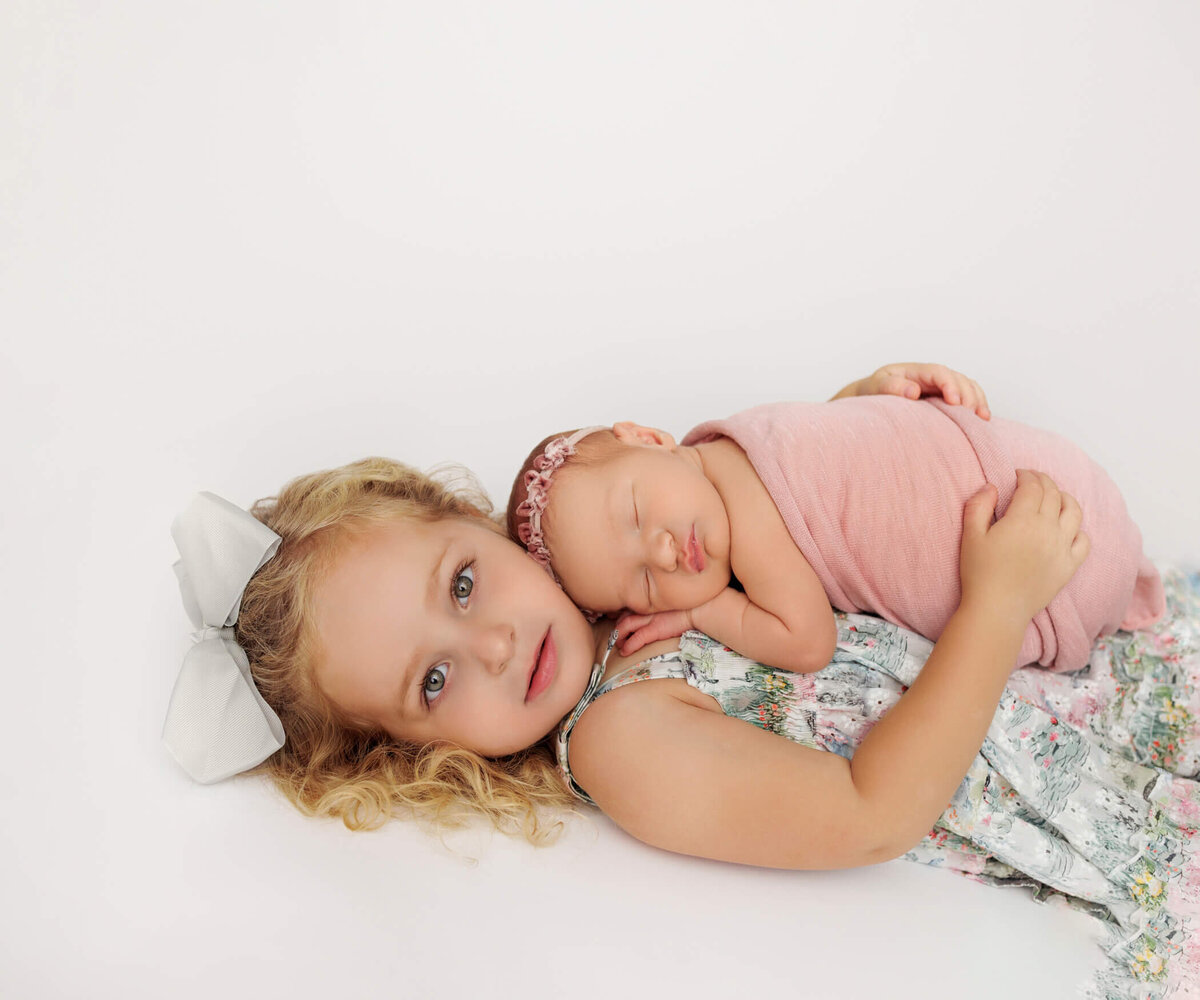 A young girl with blonde curly hair, wearing a white bow, lies on a white background, lovingly holding her newborn sibling wrapped in a soft pink swaddle. The newborn sleeps peacefully on her sister's chest.