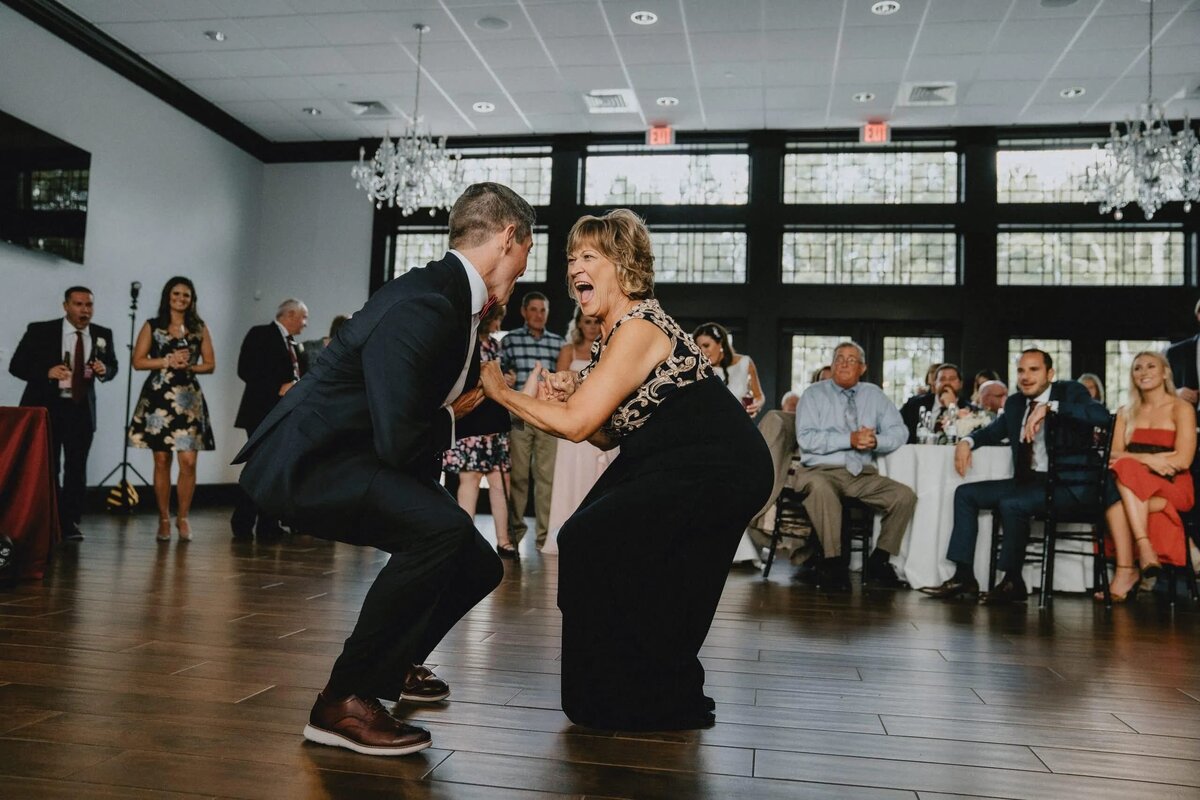 A groom and mother share a special dance, both with wide smiles, creating a joyful and memorable moment at the wedding reception.