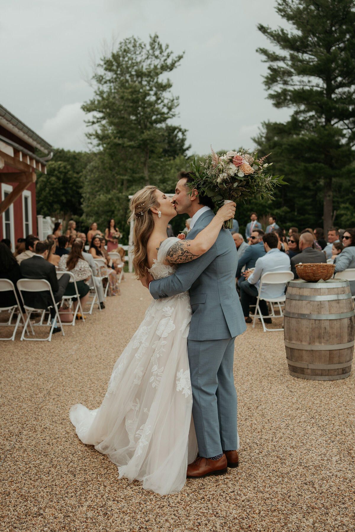 Bride and groom share a celebratory kiss after their outdoor wedding ceremony, with the bride holding a large bouquet of flowers and guests clapping in the background. The bride wears a lace gown, and the groom is in a light blue suit.