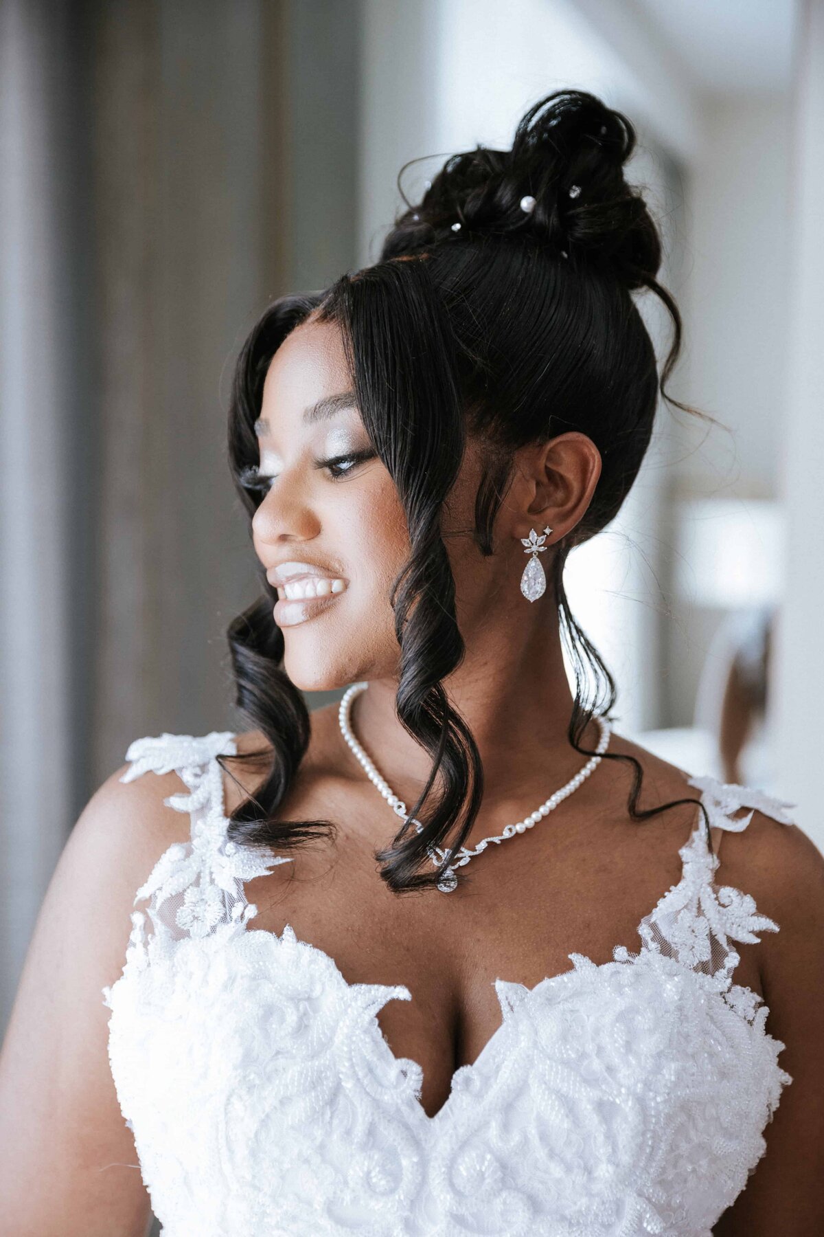 A glowing St. Augustine bride looks off-camera with a soft, joyful smile, captured in a naturally lit room. She wears a lace wedding gown with delicate floral detailing, complemented by teardrop diamond earrings and a matching necklace. Her hair is styled in a romantic updo with soft curls framing her face. Photographed by Phavy Photography in a true-to-color, editorial-meets-documentary style that captures beauty, elegance, and authentic emotion with timeless grace.