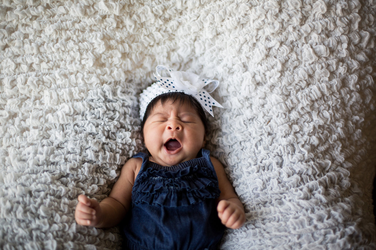 Newborn baby yawning on white flower blanket wearing blue jumper.