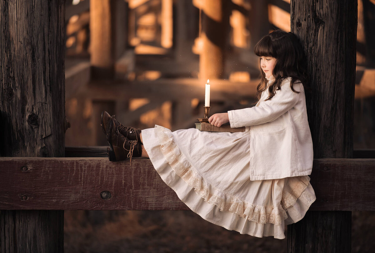 A young girl in a vintage dress sits on a bridge tressle while holding a candle on her legs. Portrait by Brittany Danielle