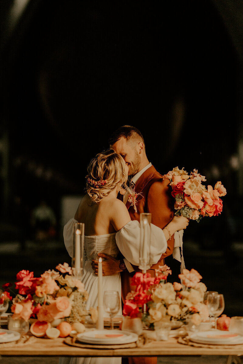 Mariés s'embrassant derrière une table en bois dressée décorée de fleurs, d'agrumes et de bougeoirs. Arrière plan noir. Photo prise par Laura Termeau, photographe professionnelle.