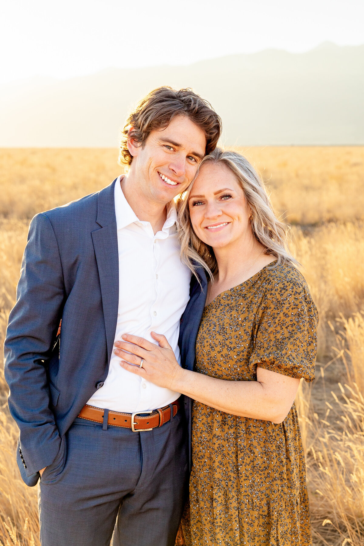A couple stands close together in a sunlit field, dressed elegantly, smiling as they pose for a timeless family portrait.