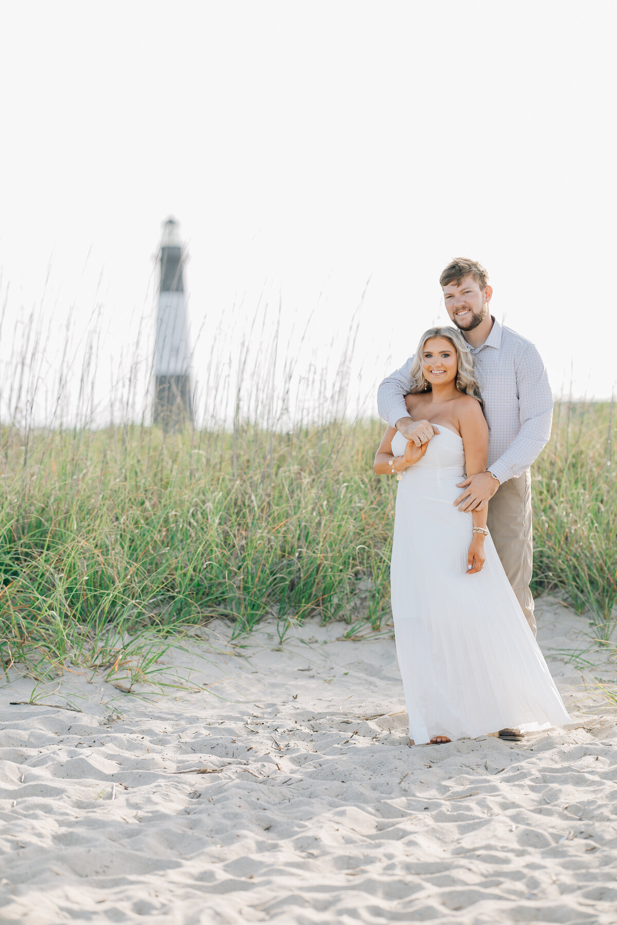 an engaged couple posing on the beach with a light house in the background