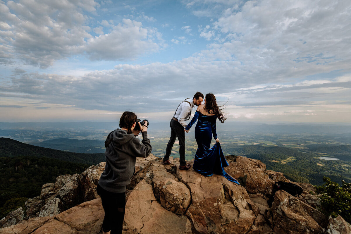 A photographer taking a picture of a couple dressed up on top of a mountain with scenic views