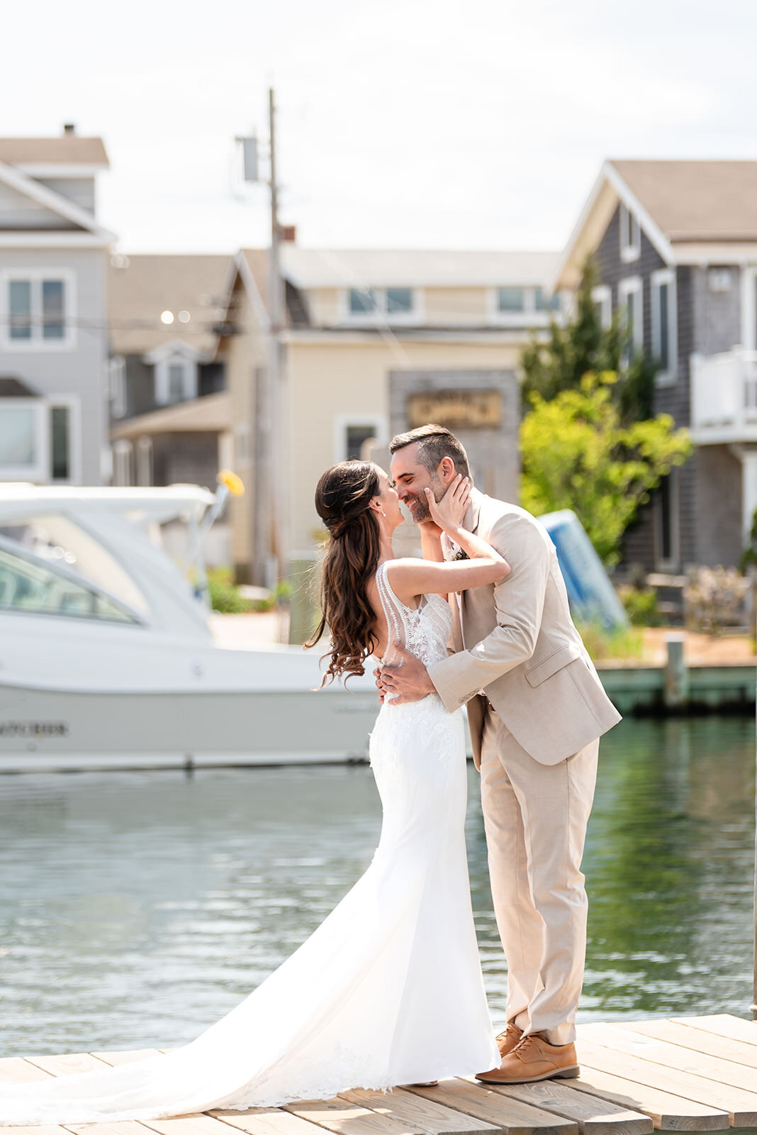 A bride and groom tenderly kissing on a sunny dock, with waterfront homes and a boat in the background.