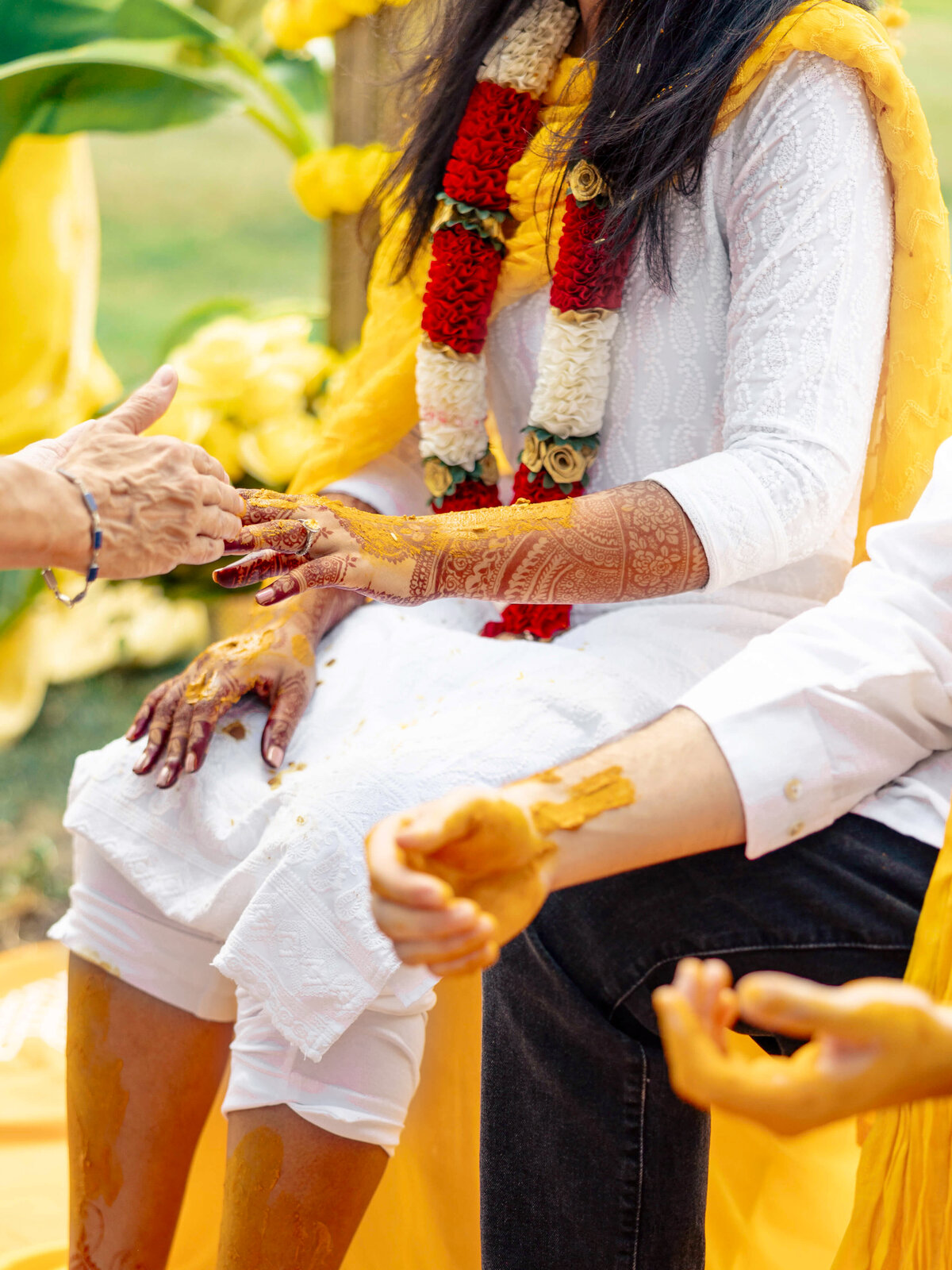 A person dressed in white with a floral garland around their neck sits while another hand extends toward them. Their hands and feet are decorated with intricate henna designs. Yellow flowers and fabric adorn the background, suggesting a festive occasion.