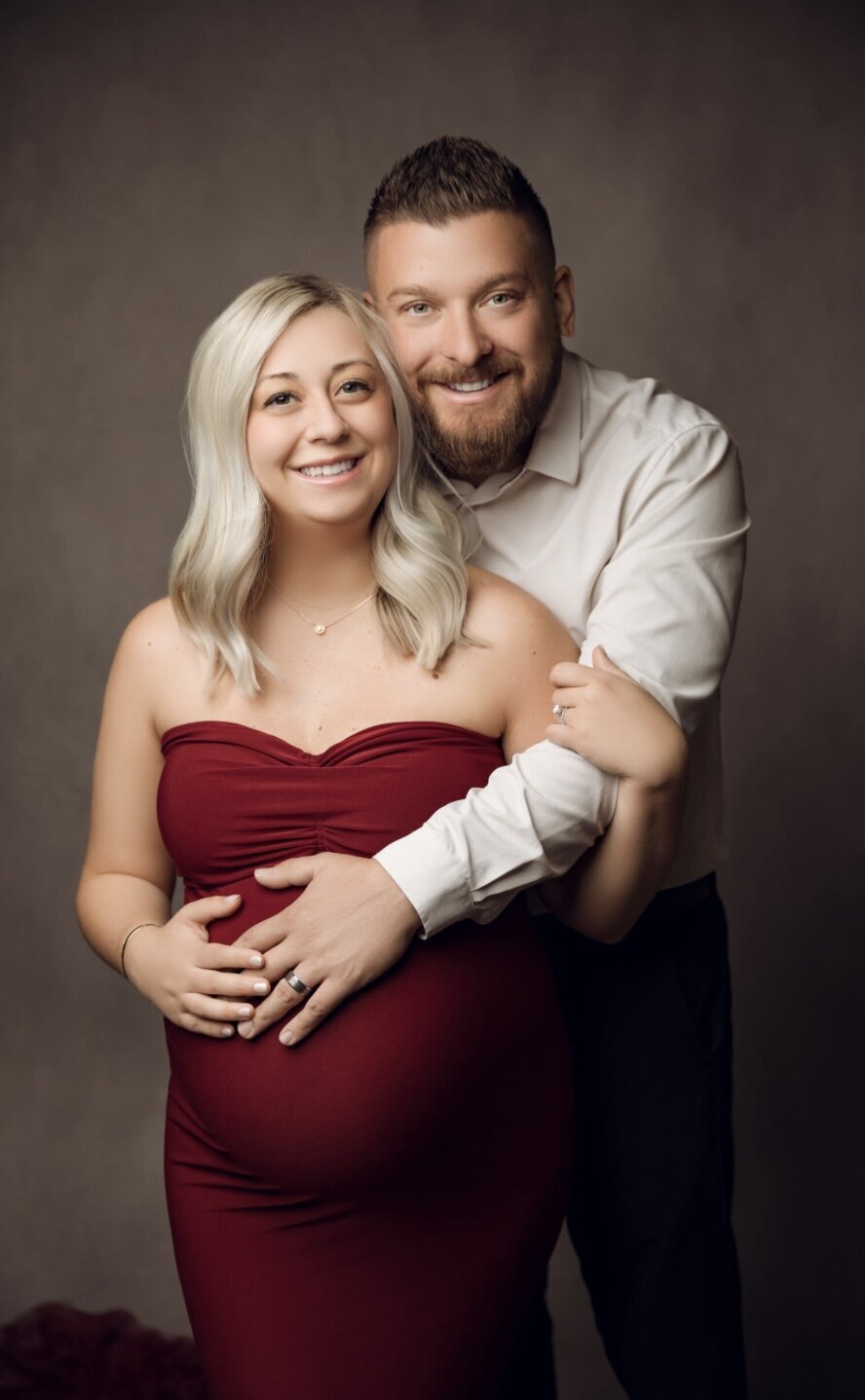 An expecting couple posing for a photo with mom wearing a red dress and dad wearing a ream dress shirt.