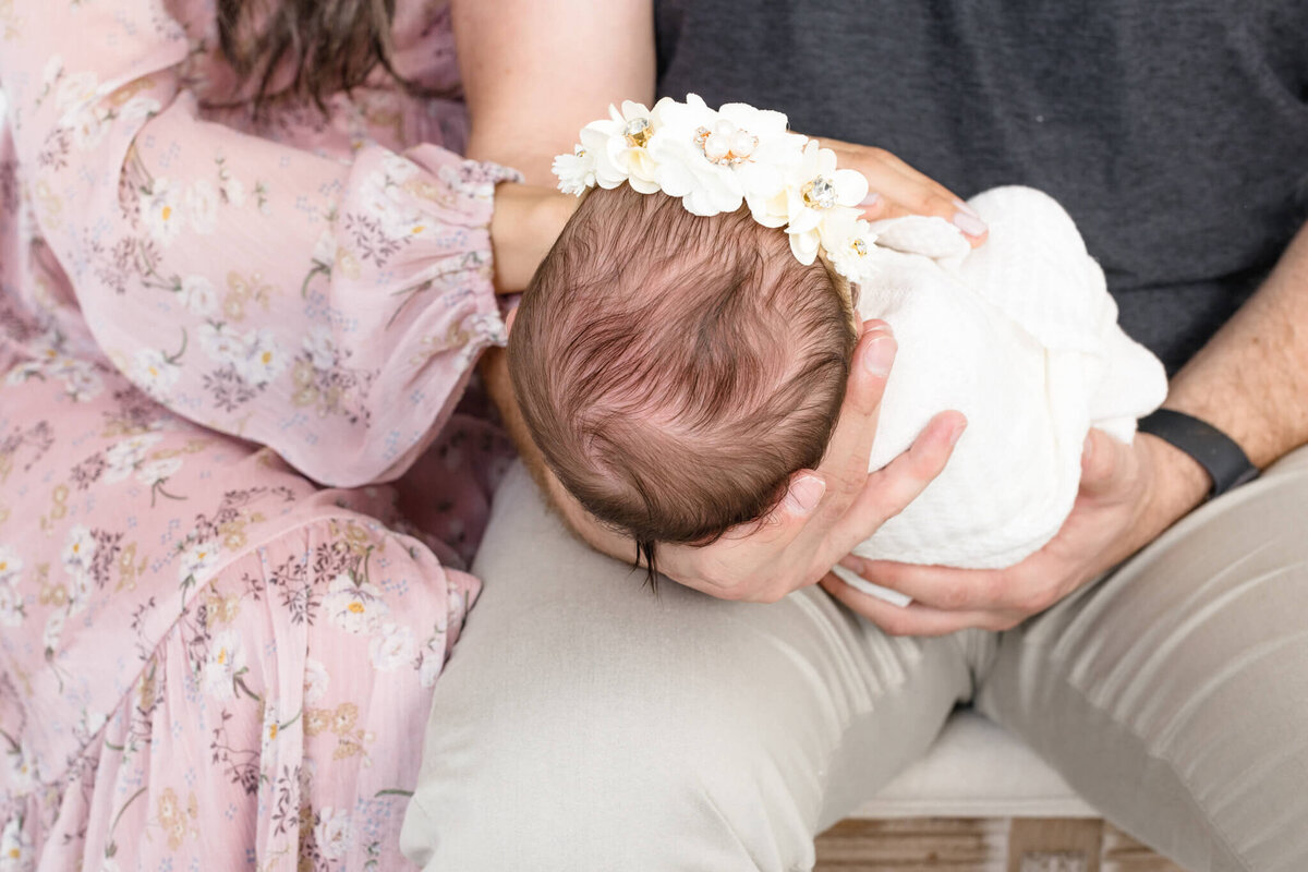 A couple holds a newborn baby dressed in white. The baby is wearing a floral headband. The father supports the babys head while seated; the mother wears a floral dress, gently touching the baby.