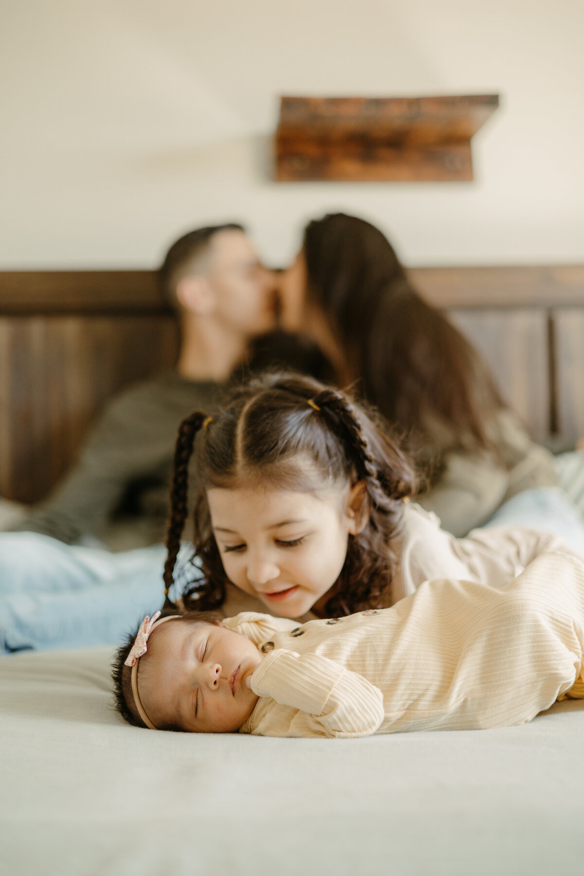 big sister looking down at newborn baby on top of bed while parents are in the background kissing during in home newborn session in Longmont Colorado
