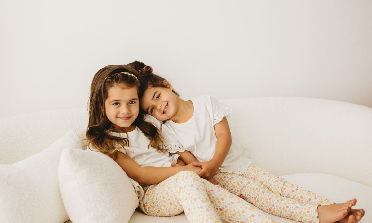 two brown hair girls sitting on a while couch and wearing white shirts as well and smiling with