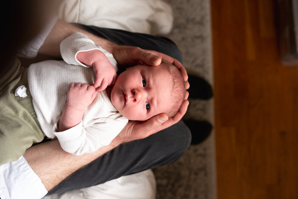 A father cradles his newborn baby in his lap, gently supporting the baby's head with his hands. The baby, dressed in a white long-sleeve onesie and olive green pants, gazes up with wide eyes. The soft, natural lighting and cozy home setting create a warm, intimate moment