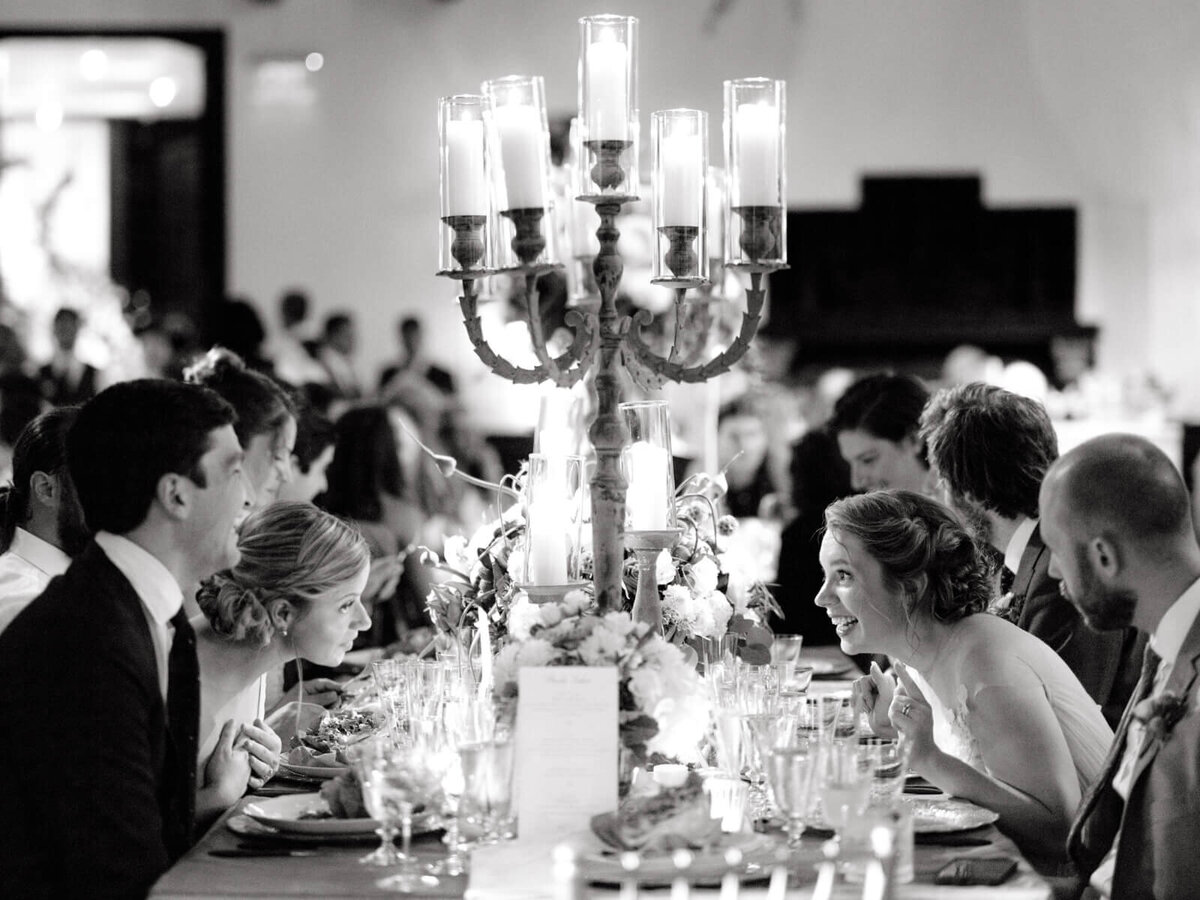 Black and white photo of a group of people, chatting with each other, on a long dining table with a large candle centerpiece.