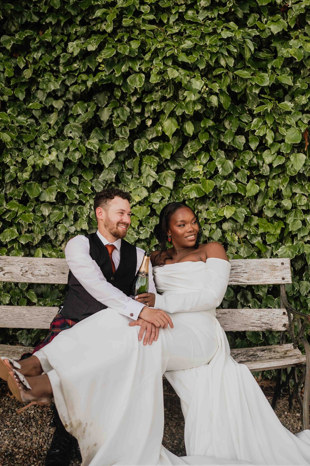 A couple sits on a wooden bench in front of a lush green ivy wall at Logie Country House in Aberdeenshire. The bride wears a flowing off-the-shoulder wedding dress, and the groom sports a dark vest and tie, smiling warmly for their photographer as they relish a joyful moment together.