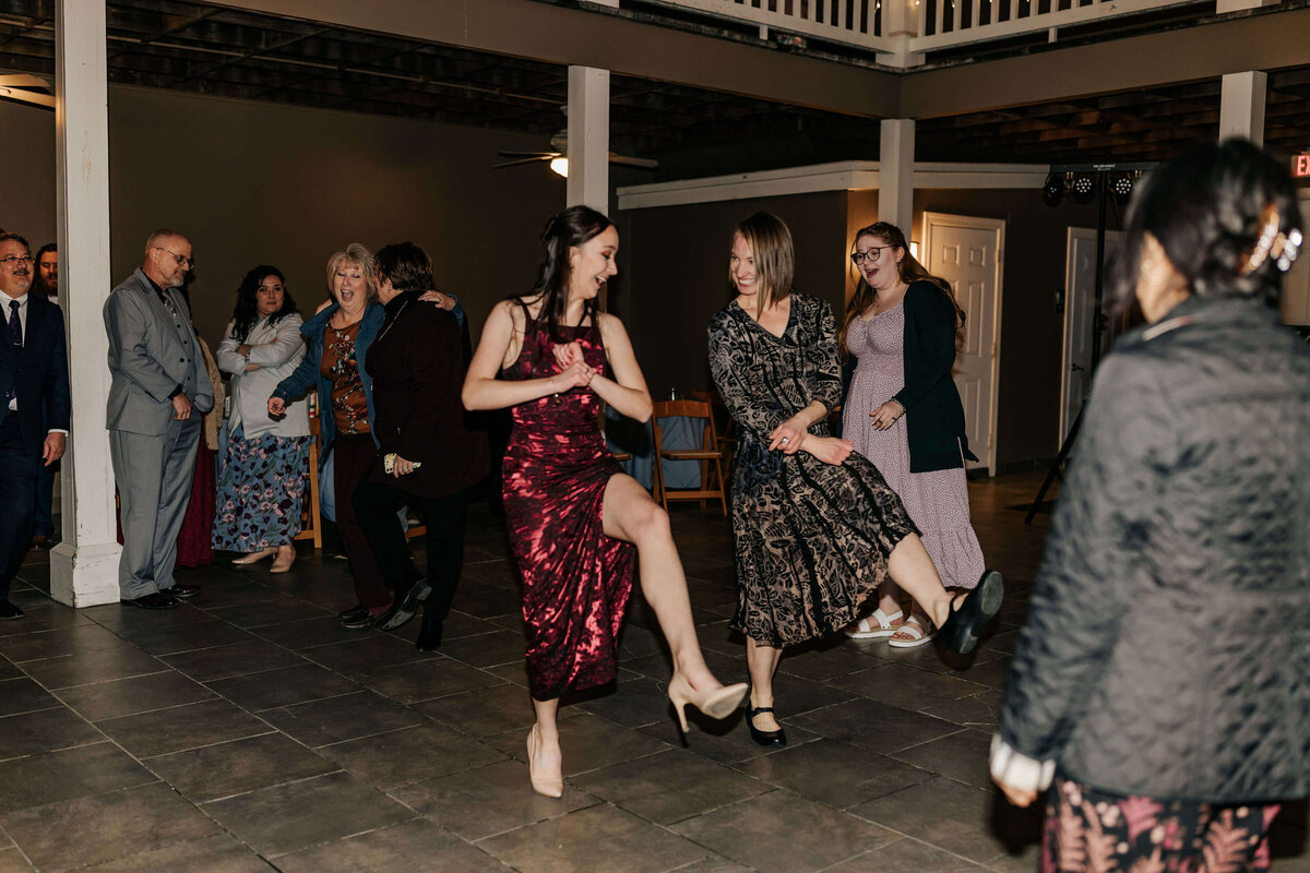A group of people at an outdoor event watch as three women in dresses enthusiastically dance and kick their legs, smiling and laughing together on the dance floor.