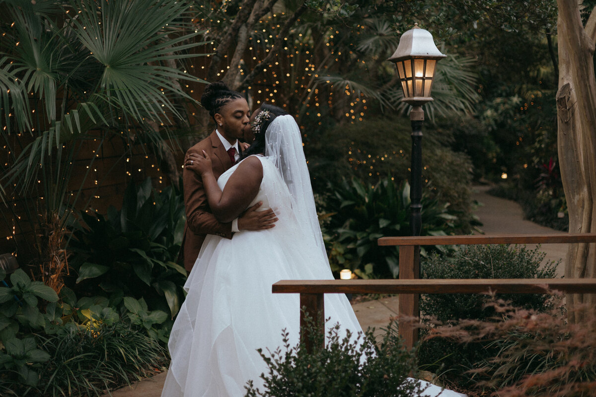 A newlywed couple shares a kiss in the gardens at the River Road and Jasmine House venue.  Twinkle lights and various greenery surrounds them.