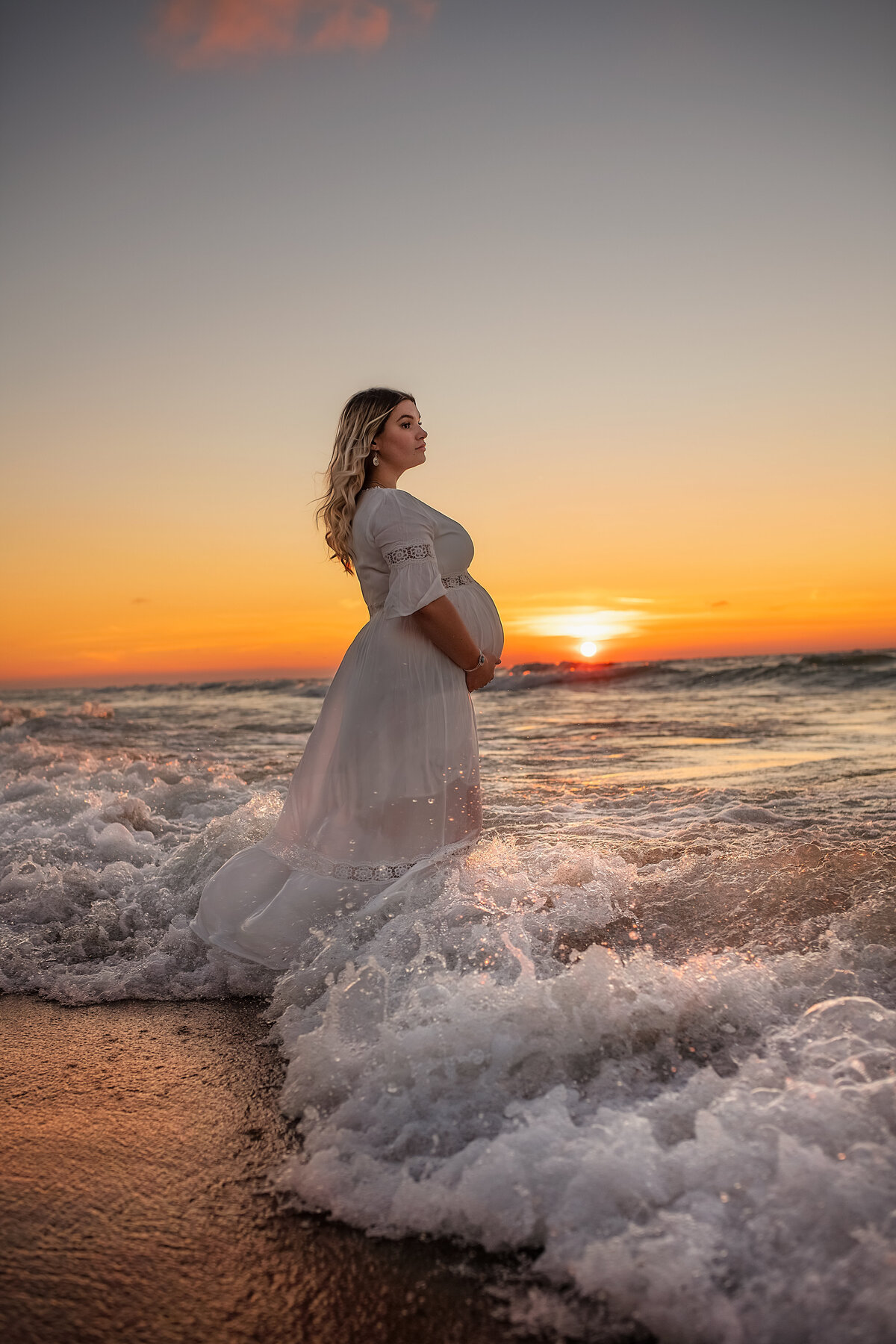 looking in to the distance while holding pregnant belly in white maternity dress during sunset at the beach