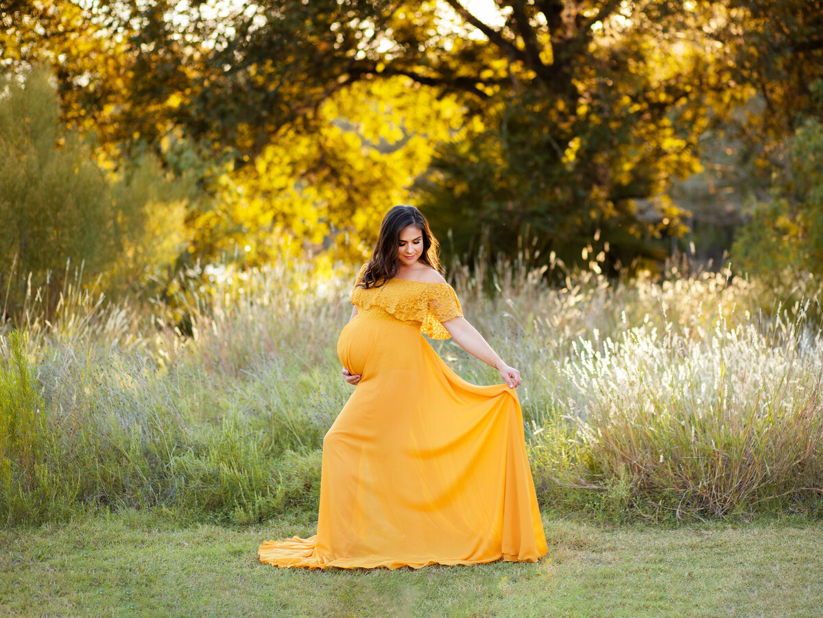 Mom in yellow dress holds dress and one hand under belly outside with sunset background